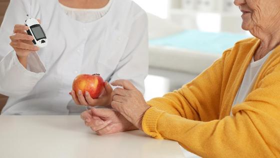 Senioren sitzt in einem Behandlungszimmer bei einer Ärztin. Sie halten ein Blutzuckermessgerät und einen Apfel in der Hand.