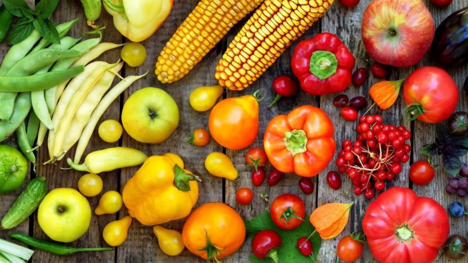 Verschiedene Obst- und Gemüsesorten liegen nach Farben sortiert nebeneinander: von grünen Bohnen und Gurken über gelbe Äpfel und Paprika hin zu roten Tomaten und schließlich blauen Beeren und Aubergine. 