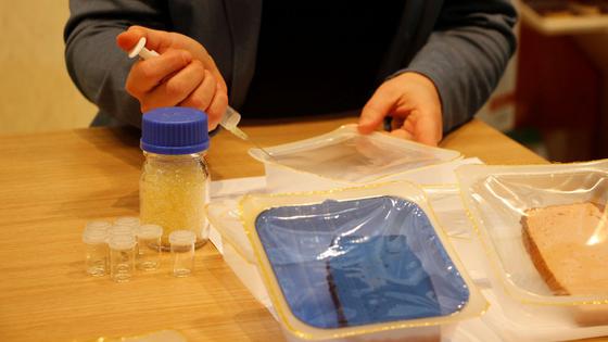 Eine Frau injiziert mit einer Spritze zu Testzwecken ein Gas in eine Fleischverpackung aus Plastik. Vor ihr liegt eine weitere Fleischverpackung mit blauem Deckel. 