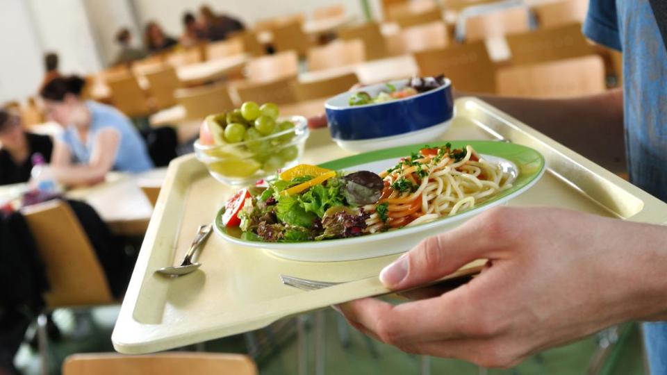 Ein Tablett mit Mittagessen, Salat und Dessert, im Hintergrund ein Kantinenraum mit Schülern