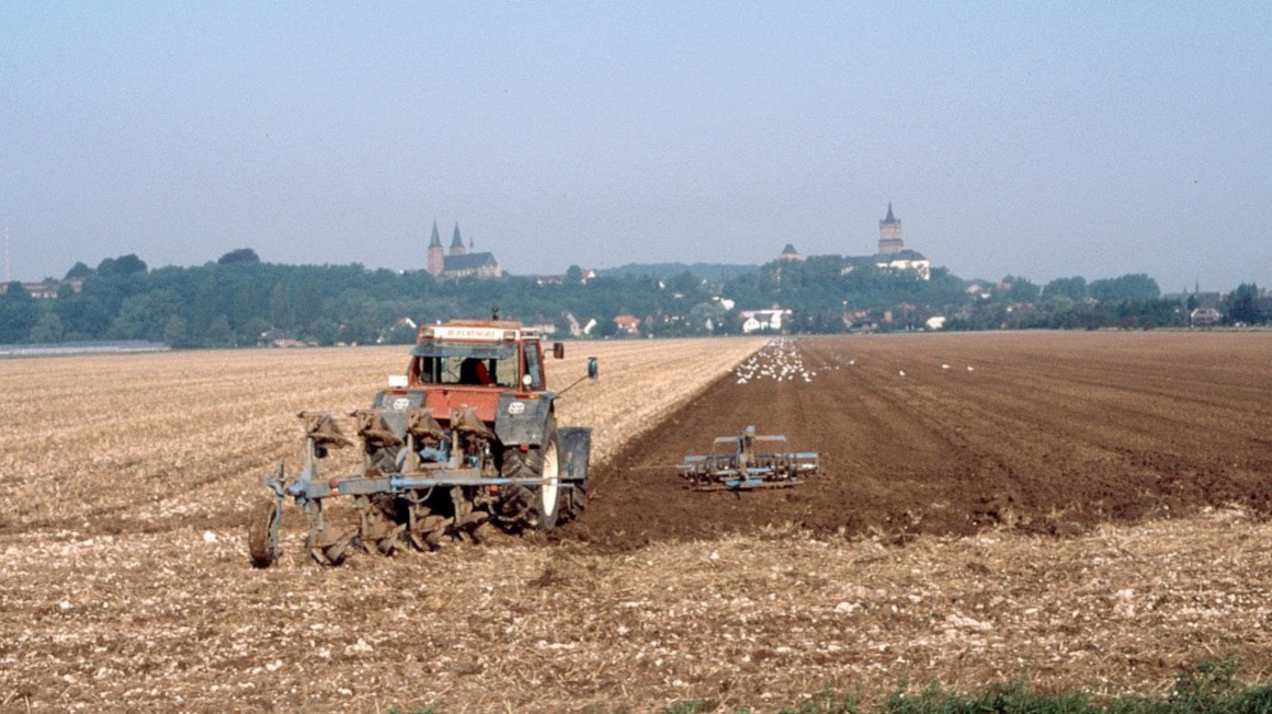 Ein Traktor pflügt ein abgeerntetes Feld, im Hintergrund eine Stadt