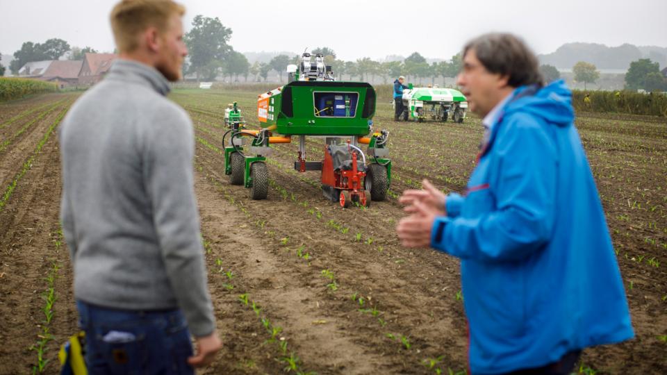Im Vordergrund zweit Wissenschaftler im Gespräch, dahinter zwei landwirtschafltiche Roboter für die Feldarbeit im Einsatz