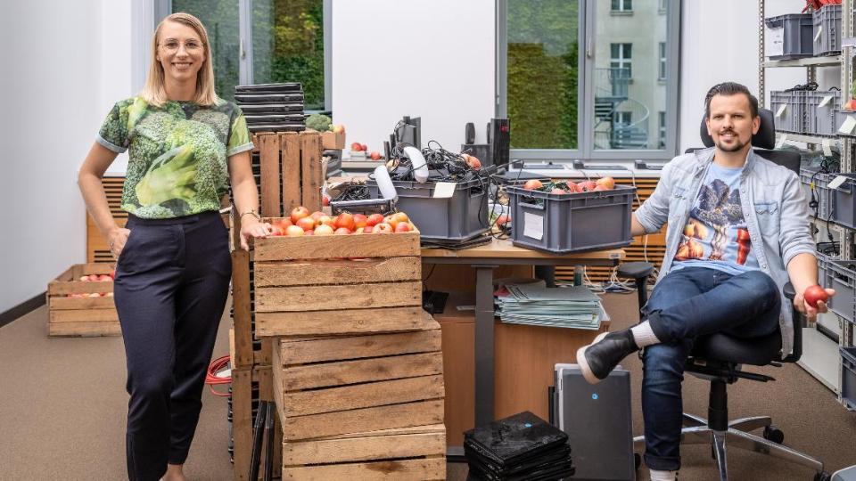 Ein Mitarbeiter und eine Mitarbeiterin des BMEL in einem Büro des Ministeriums. Um sie herum Büromöbel, Kisten mit Kabeln und Telefonen sowie Kisten voller Äpfel