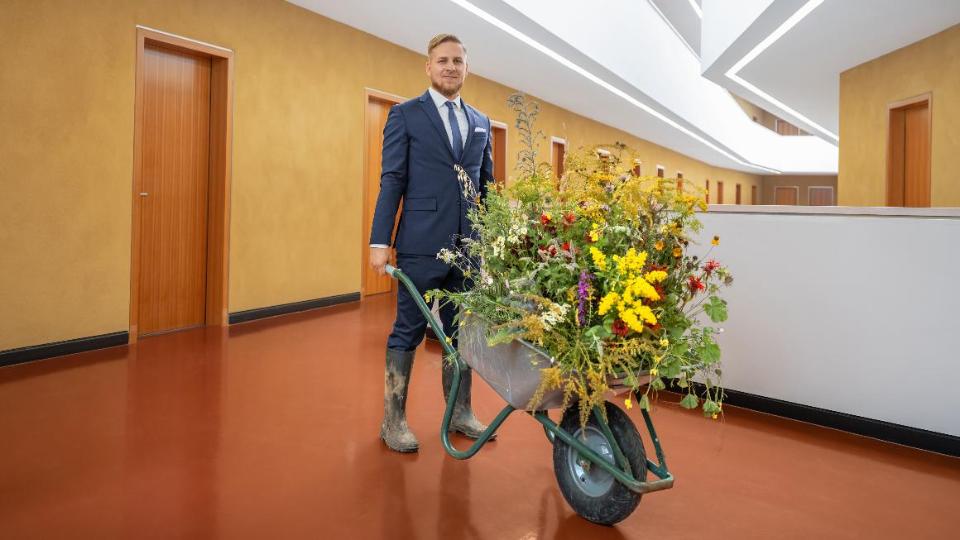 Ein Mitarbeiter des BMEL steht auf einem Flur des Ministeriums. Er trägt einen Anzug sowie matschige Gummistiefel und schiebt eine Schubkarre voller blühender Blumen.
