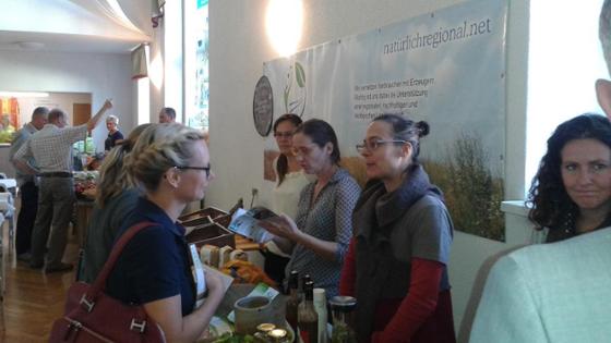 Zwei Frauen sprechen an einem Infostand mit drei Frauen von der Ökomarktgemeinschaft Thüringen/Sachsen. Auf dem Tisch zwischen ihnen stehen Flaschen, Einmachgläser, kleine Papiertüten und Holzkisten.