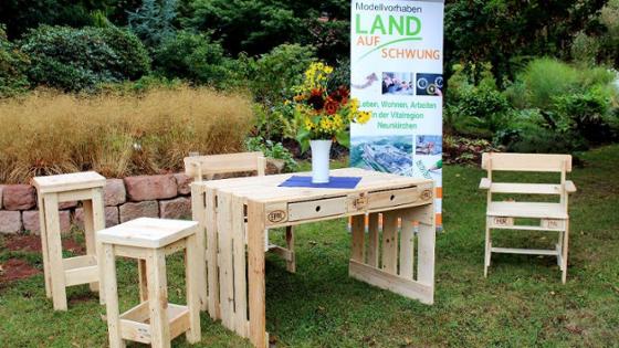 Ein Tisch und vier Hocker, gefertigt aus Europaletten, stehen auf einer Wiese in einem Garten. Auf dem Tisch ein Blumenstrauß. Zwischen zwei Hockern: Ein Roll-Up mit Informationen zu dem Projekt und dem Logo von Land(Auf)Schwung. 