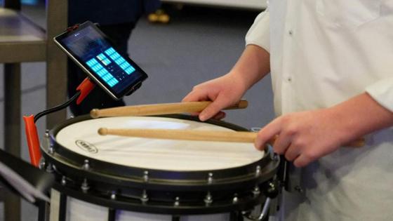 Ein Tablet mit einen Musikprogramm steckt in einer Halterung - daneben spielt eine Person auf der Trommel.