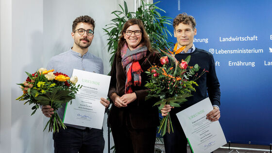 Die Parlamentarische Staatssekretärin in der Mitte, links und rechts die beiden Preisträger mit Urkunde und Blumen in der Hand.