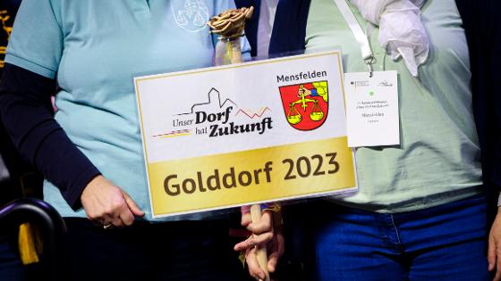 Blick auf ein Schild mit dem Wappen von Mensfelden (eine Hand, die eine Waage hält) und der Aufschrift "Unser Dorf hat Zukunft" Golddorf 2023