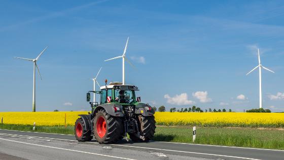 Ein Traktor fährt auf einer Straße, im Hintergrund ein Rapsfeld und Windräder.